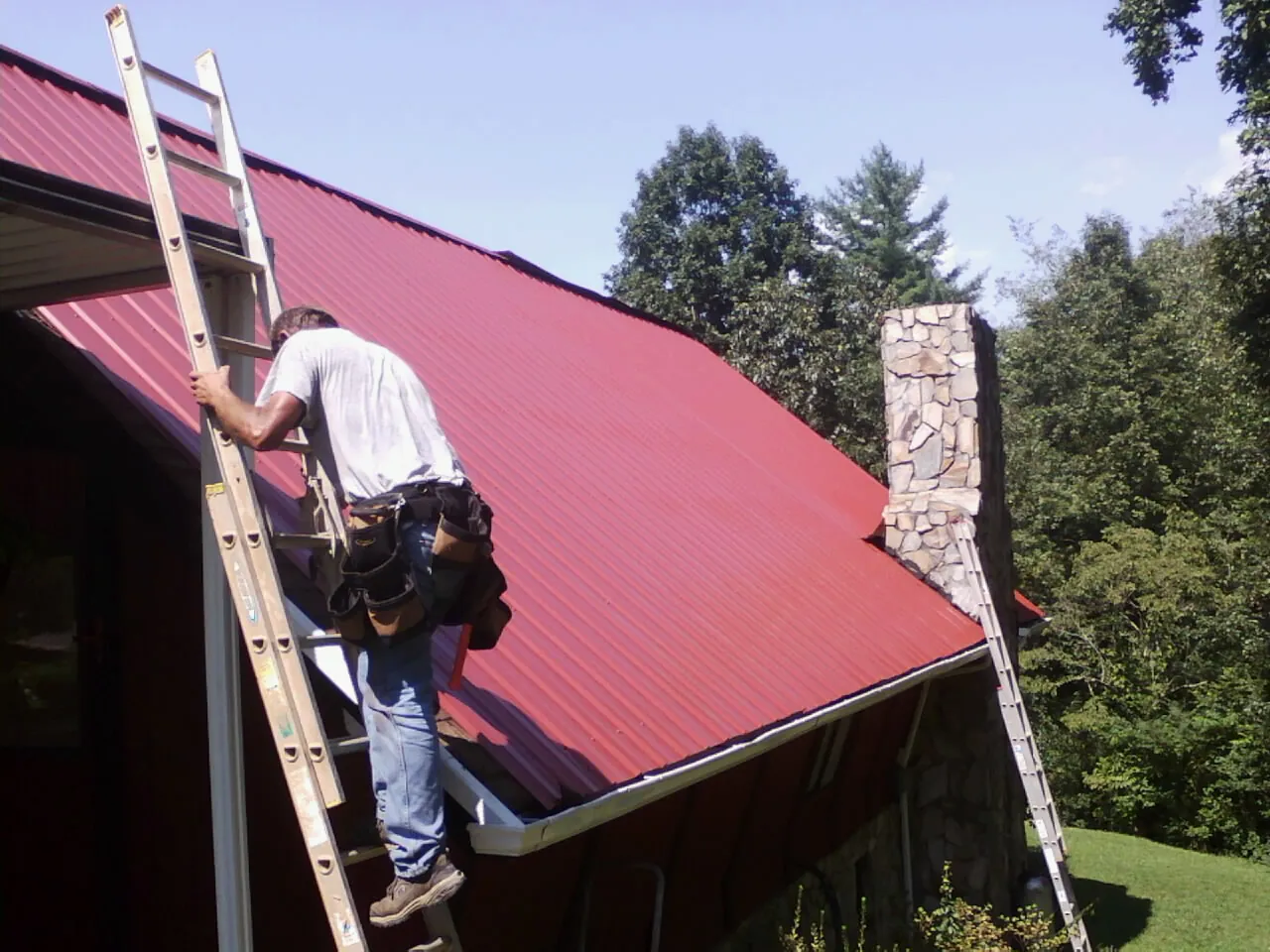 Roofer on a ladder beside a red metal roof and stone chimney — Soffit & Fascia Repair by Roofing Brothers in Menifee